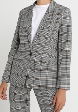Frau trägt einen grauen karierten Blazer und passende Hose über einem weißen gerippten Shirt vor neutralem Hintergrund.