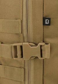 Détail d'un sac à dos en tissu beige comprenant une fermeture éclair robuste, une boucle en plastique et des sangles tissées. Étiquette de logo notable en haut à droite.
