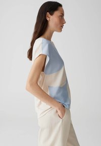 Kurzarmpullover aus einer Mischung von hellblauem und cremefarbenem Stoff, mit glatter Textur und einfachem, gebogenem Design. Trägt eine beige Hose.