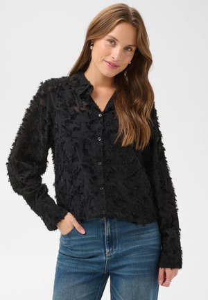 Jeune femme aux longs cheveux bruns portant une chemise noire texturée à boutons et un jean bleu, debout avec une main dans la poche devant un fond uni.
