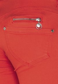 Red stretch fabric pants featuring a zippered back pocket with silver hardware and a button accent. Smooth texture with visible stitching.