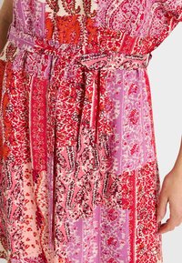 Vestido multicolorido con una mezcla de patrones rojos y rosas, diseños florales y paisley, con un cinturón de tela en la cintura y superficies texturizadas.