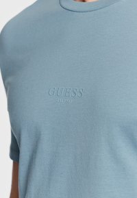 T-shirt di cotone azzurro chiaro con colletto a giro e maniche corte. Presenta il logo in rilievo 'GUESS EST. 1981' sulla parte frontale. Texture morbida.