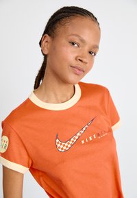 T-shirt en coton orange avec un col rond jaune, présentant un logo Nike à damier et le texte "NIKE AIR". Manches courtes avec un bord contrastant.