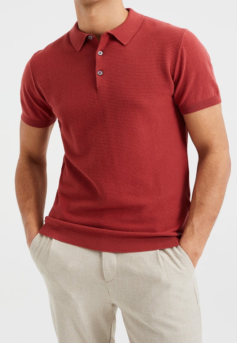 Polo en tricot rouge avec un motif texturé, manches courtes et patte de boutonnage à deux boutons. Associé à un pantalon beige clair ajusté.