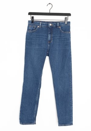 Jean en denim bleu avec poches avant et passants pour ceinture suspendu à un cintre noir sur un fond blanc.