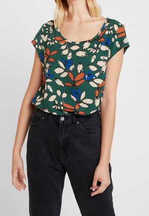 Kobieta ubrana w zieloną bluzkę z krótkim rękawem w kwiatowy wzór z kremowymi, rdzaawymi i niebieskimi liśćmi, w połączeniu z czarnymi, wysokim stanem jeansami.