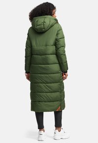 Lang groene pufferjas met capuchon, met een gequilt ontwerp, ritsdetails en zijzakken. Gekocht over zwarte leggings en witte sneakers.