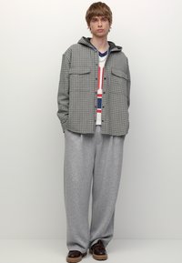 Šaljiva srajca v sivi in črni barvi, z dvema žepoma na prsih, v kombinaciji z ohlapnimi sivimi trenirkami in temno rjavimi čevlji. Preprost, priložnostni dizajn.