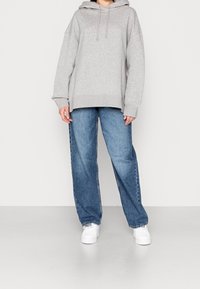 Person, der einen hellgrauen übergroßen Kapuzenpullover, lockere blaue Jeans und weiße Sneakers trägt, steht auf einem schlichten hellen Boden vor einer weißen Wand.