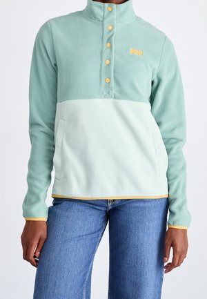 Persoon met een tweekleurige mintgroene fleecetrui met gele knopen en bies, gecombineerd met blauwe jeans, staand tegen een effen achtergrond.