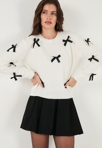 Wit gebreid sweater met zwarte strikken op de mouwen, gecombineerd met een zwarte uitlopende rok. De outfit heeft een ronde halslijn en ribbelrand.
