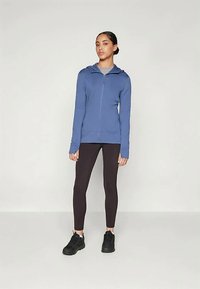 Sieviete stāv ar zilu kapuci, melnajām leggām un melnajām sporta kurpēm pret vienkāršu gaišu fona.