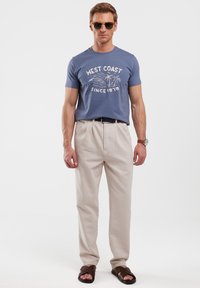 Mėlyna grafika t-shirt su užrašu "Vakarų pakrantė", smėlio spalvos plisuotos kelnės ir rudos sandalai. Aksesuarai apima saulės akinius ir laikrodį ant riešo.
