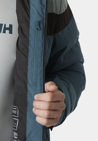 Veste matelassée bleue et noire avec une poche zippée sur le côté. Présente une texture lisse et un design en color-block.