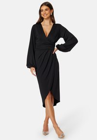 Svart omlottklänning med djup V-ringning, långa ärmar och asymmetrisk fåll. Tillverkad av slät tyg, med drapering vid midjan för en figurnära look.