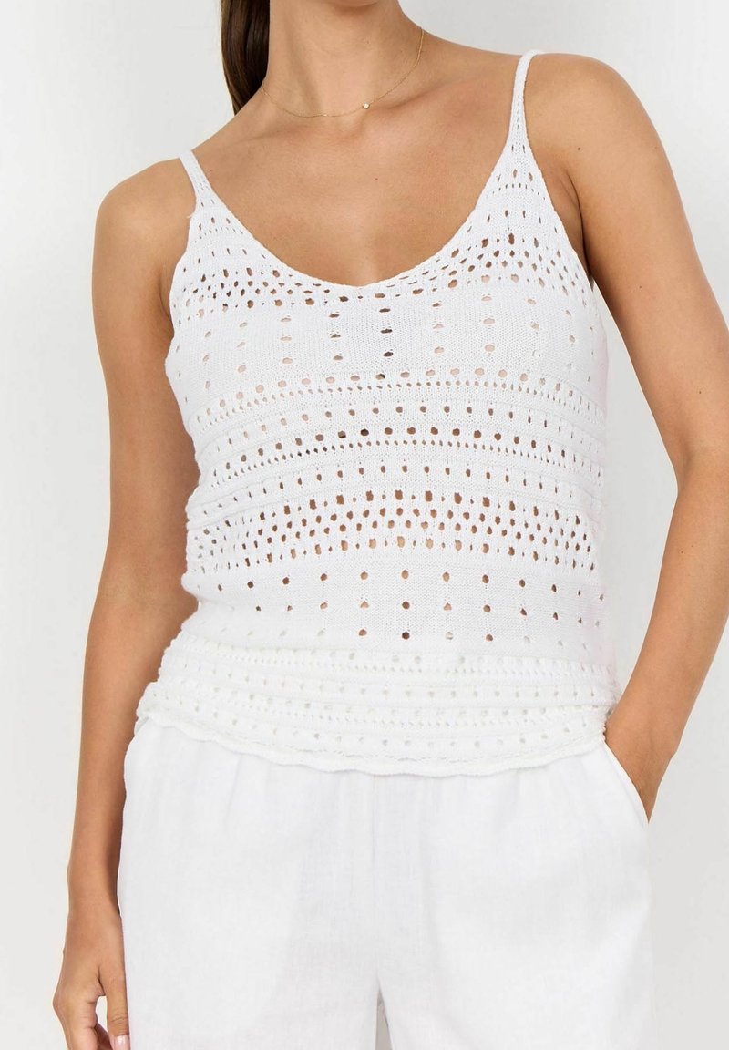 Kobieta w białej, koronkowej, bezrękawnikowej bluzce z cienkimi ramiączkami i białych spodniach, z ręką w kieszeni, na jednolitym tle.