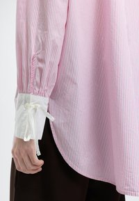 Μανίκι ροζ και λευκής ριγέ μπλούζας με λευκή μανσέτα που διαθέτει κουμπί και δεμένη υφασμάτινη φιόγκο, φορεμένο με σκούρο παντελόνι.