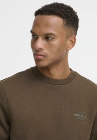 Sudadera marrón con cuello redondo acanalado. Presenta un pequeño detalle de texto impreso en el lado izquierdo del pecho. Textura suave, ajuste informal.