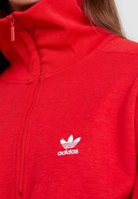 Röd dragkedjas blus i mjukt tyg. Har en hög krage och en vit Adidas-logotyp på vänster sida. Slät textur.