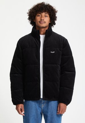 Homme aux cheveux bouclés portant une doudoune en velours côtelé noire par-dessus une chemise blanche et un jean bleu, debout devant un fond uni.