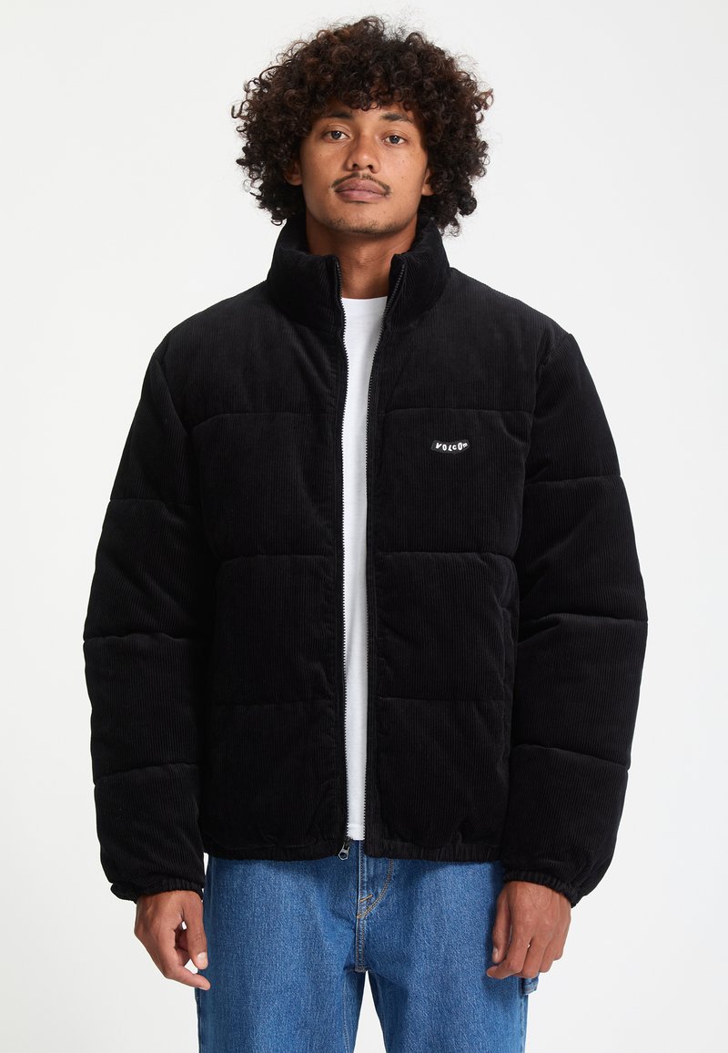 Homme aux cheveux bouclés portant une doudoune en velours côtelé noire par-dessus une chemise blanche et un jean bleu, debout devant un fond uni.