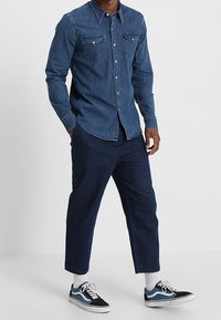 Denim-paita, jossa on suunniteltu istuvuus, tummansininen väri, kaksi rintataskua ja nappilistallinen etuosa, yhdistettynä tummansinisiin capri-housuihin ja lenkkitossuihin.