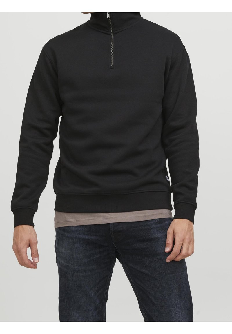 Sort lynlås-sweatshirt med høj krave, ribbet manchetter og kant samt en tætsiddende pasform, båret over en beige skjorte. Jeansene er mørkeblå.