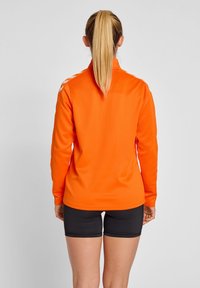 Langærmet orange atletisk top med høj krave og hvide skulderstriber, parret med sorte shorts. Glat tekstur, atletisk pasform.