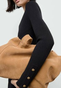 Kvinna i svart ribbad långärmad topp som håller en stor sandfärgad mockaväska med utskurna handtag framför en neutral bakgrund.