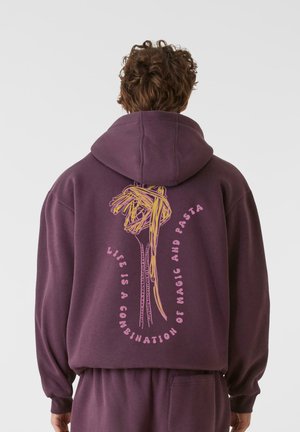 Lilla hettegenser med et grafisk motiv av spaghetti og tekst i rosa som sier "Livet er en kombinasjon av magi og pasta." Myk stoff, avslappet passform.