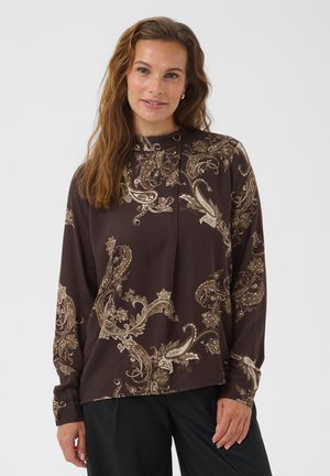 Femme aux longs cheveux bruns portant un chemisier marron avec des motifs cachemire beiges et un pantalon noir, debout devant un fond clair uni.