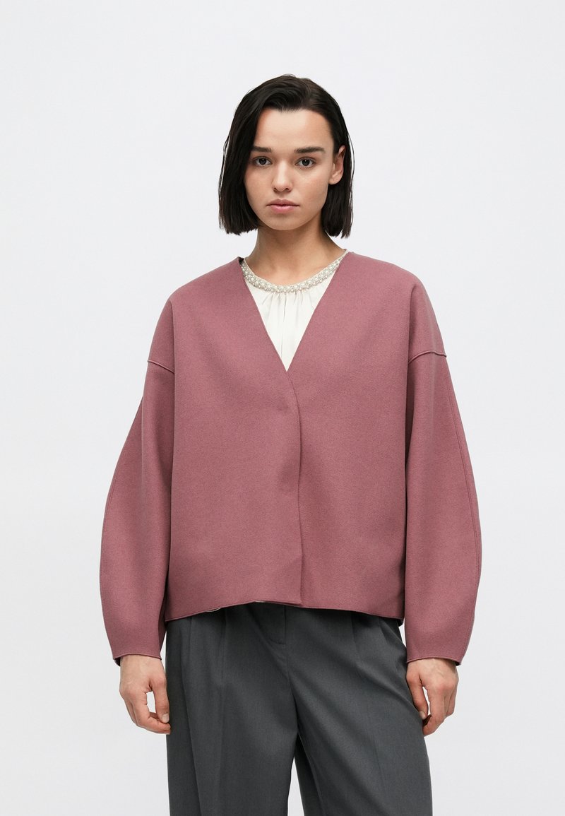 Oversized jacka i dammrosa med v-ringning och sänkta axlar, tillverkad av ett mjukt tyg, med en minimalistisk design och rena linjer.