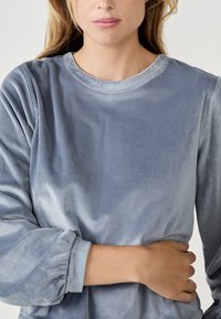 Top de pana acanalada azul claro con escote redondo y puños de manga fruncidos. Textura suave, diseño ajustado y un brillo sutil.