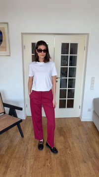 Žena nosiace čierne slnečné okuliare, biele tričko Calvin Klein, magentové nohavice a čierne mokasíny, stojaca v miestnosti s drevenou podlahou a sklenenými dverami.