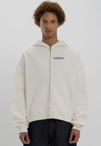 Felpa con zip color crema in tessuto morbido con tasca frontale; presenta un logo sul lato sinistro del petto e un cappuccio regolabile con cordino.