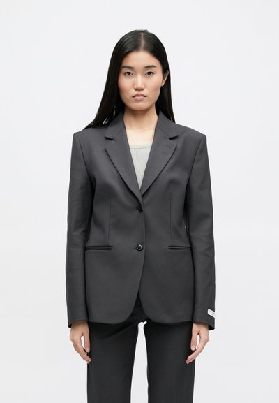 Σκούρο γκρι σακάκι προσαρμοσμένο με δομημένο σχέδιο, δύο κουμπιά και διακριτές ραφές. Το ύφασμα φαίνεται λείο, συνδυασμένο με κομψά παντελόνια.
