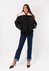 Giubbotto puffer nero con colletto in pelliccia sintetica bianca, design trapuntato, orlo a coste e vestibilità oversize, abbinato a jeans blu e tacchi neri.