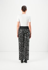 Sieviete stāv ar muguru pret skatītāju, ģērbusies baltā īsajā piedurknē un melnās platās biksēs ar sudraba mirdzošām detaļām, matiem savīti bunā.