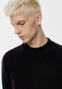 Maglione nero a coste con colletto alto e rotondo. Il tessuto sembra morbido e strutturato, indossato da un modello con capelli biondo chiaro.