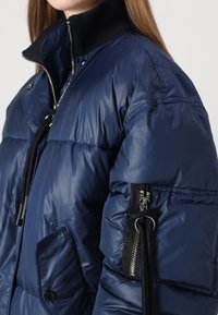 Jūras spēku puffer jaka ar augstu apkakli, ar kabatu ar rāvējslēdzēju uz piedurknes, spīdīga tekstūra un šūtu kviltēto dizainu.