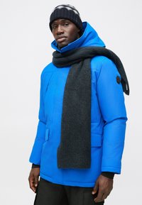 Bright blue parka jacket with a high collar, paired with a dark gray knitted scarf. The design features front pockets and a logo badge on the sleeve.