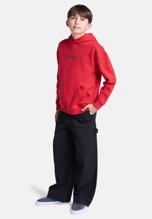 Felpa rossa con cappuccio e tasca frontale, con testo logo nero. Indossata con pantaloni neri larghi e scarpe grigie.