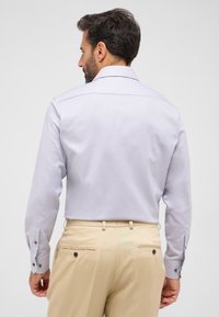 Hellpurpurnes, gemustertes Langarmhemd mit geknöpften Manschetten, kombiniert mit beigen Hosen mit Gesäßtaschen und Knopfakzenten.