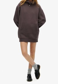 Överdimensionerad mörk mauve hoodie med en framficka, snörning i huvan, ribbad nederkant, bärs över en kort längd, tillsammans med grå strumpor och svarta skor.