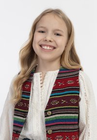 Stickad väst med röda, blå och gröna ränder, som har cirkulära mönster och metallknappar, bärs över en texturerad vit blus.