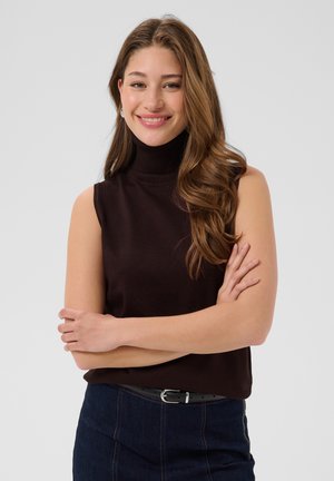 Joven mujer con cabello largo y ondulado, que lleva un suéter negro sin mangas de cuello alto y vaqueros oscuros, de pie con los brazos cruzados y sonriendo.