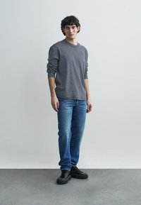 Langärmliges, gestreiftes graues und schwarzes Hemd, blaue Jeans und schwarze Sneakers, stehend auf einem grauen Boden vor einem schlichten Hintergrund.