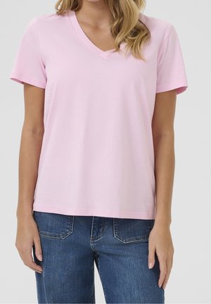 Nainen pukeutuneena vaaleanpunaiseen V-kaula-aukkoinen lyhythihainen T-paitaan ja sinisiin farkkuihin yksiväristä taustaa vasten.