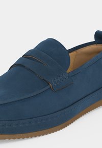 Mocassin en daim bleu marine avec bride penny, doublure intérieure beige et semelle en caoutchouc brun texturée, présenté en gros plan.